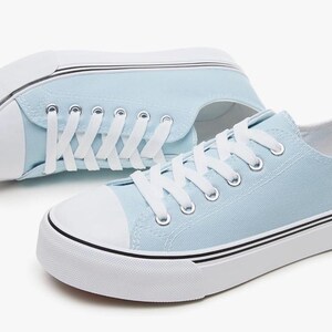 May include: A pair of light blue canvas sneakers featuring white laces, white rubber toe caps, and thick white soles with thin black horizontal stripes running along the sides. The shoes have silver-toned metal eyelets for the white laces.