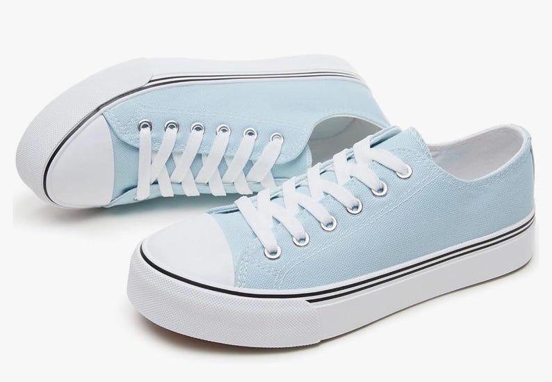 May include: A pair of light blue canvas sneakers featuring white laces, white rubber toe caps, and thick white soles with thin black horizontal stripes running along the sides. The shoes have silver-toned metal eyelets for the white laces.