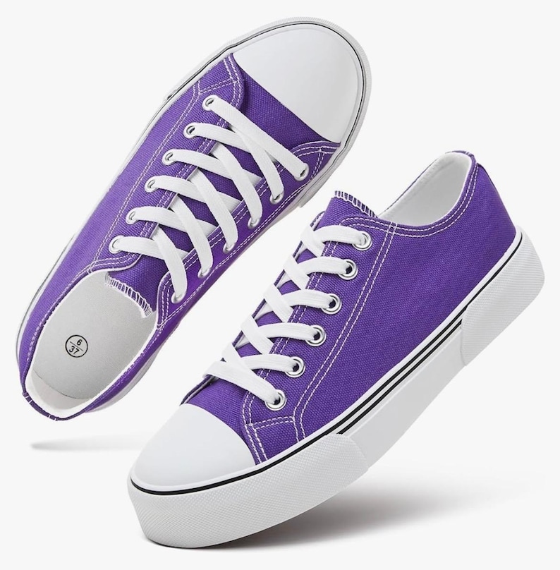 May include: A pair of purple canvas sneakers featuring white laces, white rubber toe caps, and thick white soles with thin black stripes. The interior of the shoe shows a size label with the numbers 6 and 37 printed on a white background.