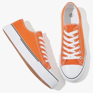 May include: A pair of low-top canvas sneakers in a vibrant orange color. The shoes feature white laces, white rubber toe caps, and thick white soles with a thin black stripe detail. The interior sole of the right shoe displays the size 6 and 37.