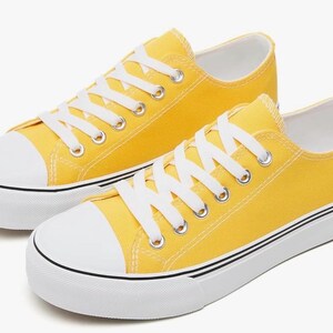 May include: A pair of low-top sneakers in a bright yellow canvas material. The shoes feature white rubber toe caps, white laces, and thick white soles with a thin black stripe running along the side of the midsole. The design is simple and clean.