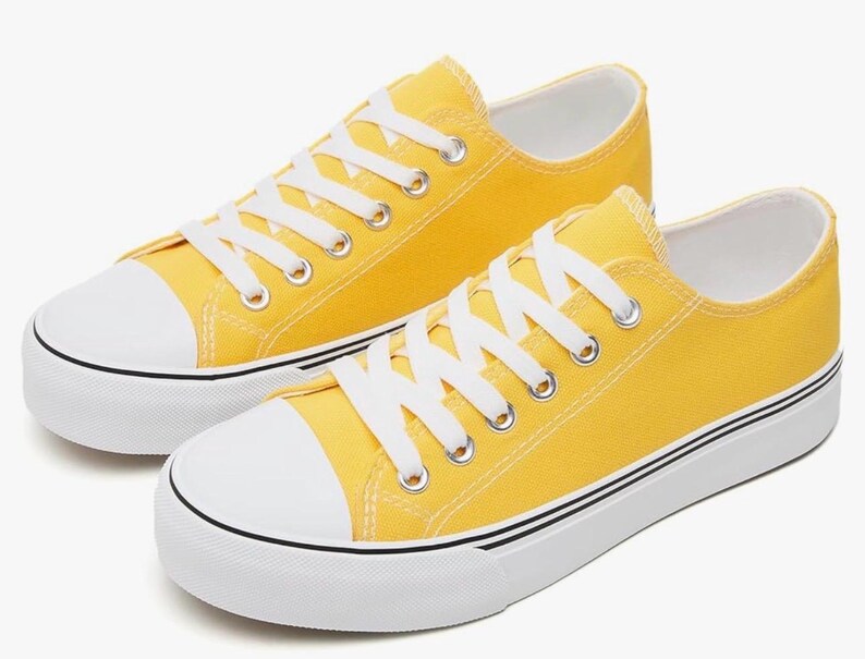 May include: A pair of low-top sneakers in a bright yellow canvas material. The shoes feature white rubber toe caps, white laces, and thick white soles with a thin black stripe running along the side of the midsole. The design is simple and clean.
