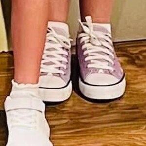 May include: A pair of light purple canvas sneakers with white laces, white rubber toe caps, and white soles with a thin black stripe. Next to them is a white sneaker with a hook-and-loop strap closure and a ruffled sock at the ankle.