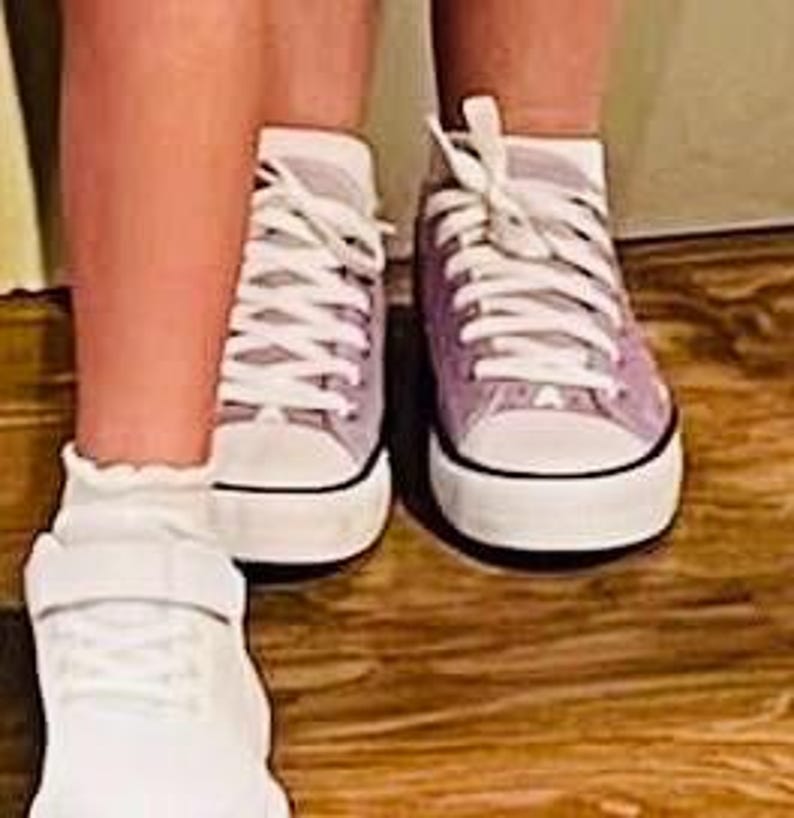 May include: A pair of light purple canvas sneakers with white laces, white rubber toe caps, and white soles with a thin black stripe. Next to them is a white sneaker with a hook-and-loop strap closure and a ruffled sock at the ankle.