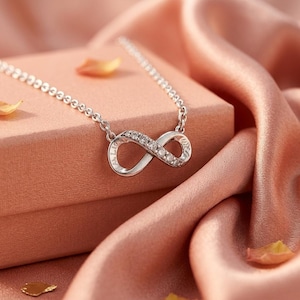 May include: A silver-toned infinity symbol pendant necklace featuring a delicate chain and a central infinity loop charm. The charm is embellished with small, clear, sparkling stones along one side and includes engraved text along the metal surface.