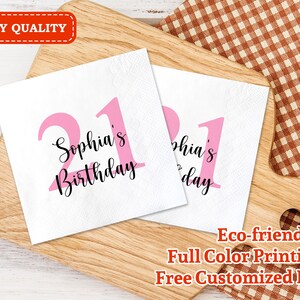 May include: Two pink and white paper napkins with the text "Sophia's Birthday" and the number "21" printed on them. The napkins are on a wooden surface with a checkered fabric in the background.