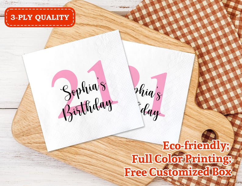 May include: Two pink and white paper napkins with the text "Sophia's Birthday" and the number "21" printed on them. The napkins are on a wooden surface with a checkered fabric in the background.