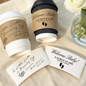 Puede incluir: Dos tazas de café de papel blanco con mangas marrones y tapas negras, y dos mangas a juego. Las mangas tienen el texto "Little Latte is Brewing" y "Welcome Baby!" con huellas de bebé.