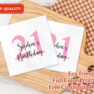 May include: Two pink and white paper napkins with the text "Sophia's Birthday" and the number "21" printed on them. The napkins are on a wooden surface with a checkered fabric in the background.