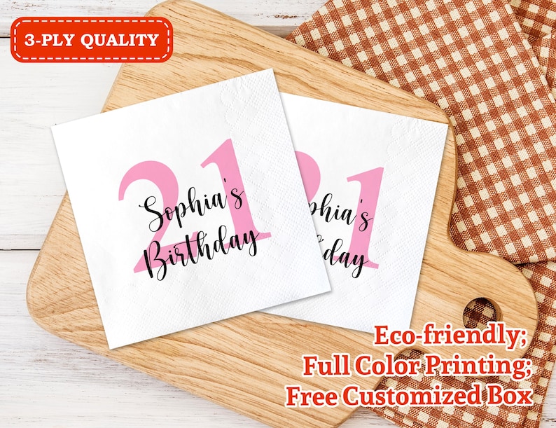 May include: Two pink and white paper napkins with the text "Sophia's Birthday" and the number "21" printed on them. The napkins are on a wooden surface with a checkered fabric in the background.