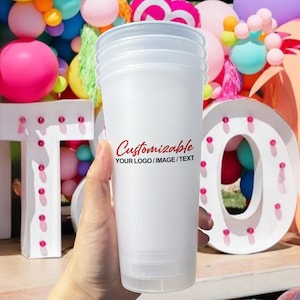 Puede incluir: Una pila de cuatro vasos de plástico blanco con tapas. Los vasos tienen un aspecto esmerilado y presentan la palabra "Customizable" en escritura roja, junto con el texto "YOUR LOGO / IMAGE / TEXT". El fondo incluye globos coloridos y una gran letra "O" iluminada.