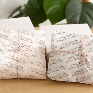 Puede incluir: Dos regalos envueltos sobre una superficie de madera. Los regalos están envueltos en papel blanco impreso con el texto "YOUR CUSTOM LOGO/IMAGE/TEXT" en un patrón repetido. Cada regalo está atado con hilo rojo y blanco.
