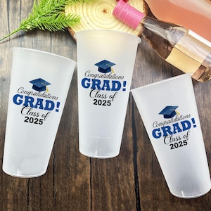 以下が含まれることがあります： 青と黒で「Congratulations GRAD! Class of 2025」の文字が入った白いプラスチックカップ3つ。各カップには卒業帽のグラフィックが描かれています。カップは木製の表面に置かれ、背景にはワインボトルがあります。