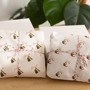 Peut inclure: Deux cadeaux emballés dans des boîtes blanches. Les cadeaux sont enveloppés dans du papier blanc avec des motifs répétés d'un elfe de Noël et le mot "Christmas" en rouge. Ils sont attachés avec de la ficelle rouge et blanche.