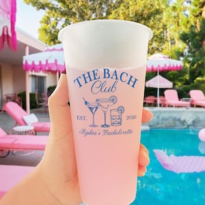 May include: A clear plastic cup filled with a pink drink, with the blue text "The Bach Club" and cocktail illustrations. The cup is held up, with a pool and pink sun loungers in the background. The text also reads "Sophia's Bachelorette".