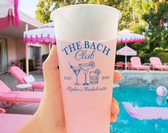 May include: A clear plastic cup filled with a pink beverage, featuring the text "The Bach Club" in blue, along with cocktail illustrations and "Sophia's Bachelorette" below. The cup is held in front of a pool and lounge chairs.