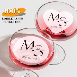 Puede incluir: Dos copas de vino llenas de líquido rosa, cada una con una pegatina en forma de corazón. Las pegatinas muestran las iniciales "MS", el nombre "The Johnsons" y la fecha "12 de junio de 2025". También incluye el texto "100% EDIBLE PAPER EDIBLE INK".