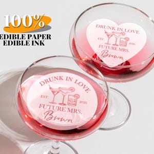 Puede incluir: Dos copas de vino llenas de líquido rosa, cada una con un diseño de papel comestible en forma de corazón. El diseño dice "Drunk in Love", "Future Mrs. Brown" y "2026". También se ve el texto "100% Papel Comestible Tinta Comestible".