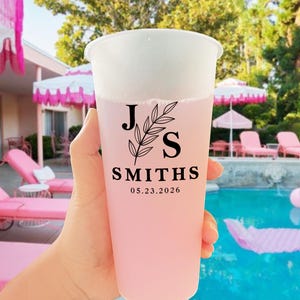 Puede incluir: Un vaso de plástico transparente lleno de una bebida rosa, sostenido por una mano. El vaso tiene una tapa blanca y un diseño floral negro con las iniciales "JS", el nombre "SMITHS" y la fecha "05.23.2026".