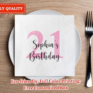 May include: A white napkin with pink numbers 21 and the text "Sophia's Birthday" printed on it. The napkin is on a wooden table with a silver fork.