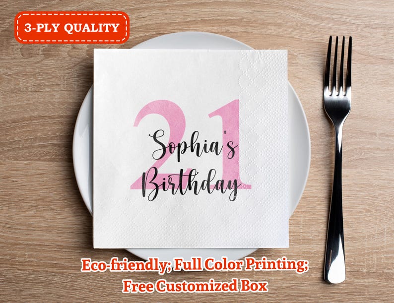 May include: A white napkin with pink numbers 21 and the text "Sophia's Birthday" printed on it. The napkin is on a wooden table with a silver fork.