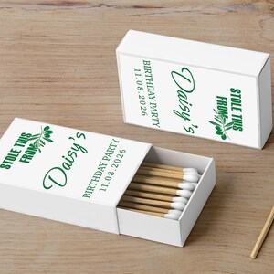 May include: White matchboxes with green text that reads "Daisy's Birthday Party 11.08.2026" and "Stole this from". The matchboxes are open, revealing wooden matches with white tips. One match is separate.