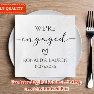 Puede incluir: Una servilleta de papel blanca con las palabras "WE'RE engaged" en escritura negra, un símbolo de corazón, nombres y una fecha. La servilleta está sobre un plato blanco con un tenedor plateado a la derecha. El texto "3-PLY QUALITY" está en un recuadro rojo.
