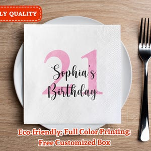 May include: A white napkin with pink numbers 21 and the text "Sophia's Birthday" printed on it. The napkin is on a wooden table with a silver fork.