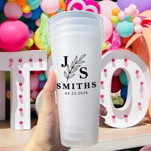 May include: A stack of white plastic cups with a black floral design and the text "J S SMITHS 05.23.2026". The cups are held in front of a colorful party backdrop with balloons and illuminated letters.