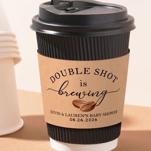 Puede incluir: Una taza de café negra con una funda de papel marrón que dice "DOUBLE SHOT is brewing" con ilustraciones de granos de café. La taza es para un baby shower el 26 de agosto de 2026. Una pila de tazas blancas está en el fondo.