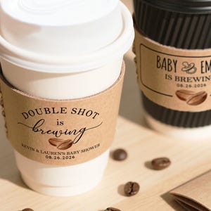 May include: Two coffee cups with lids and brown sleeves. One cup has the text "DOUBLE SHOT is brewing" and the other says "BABY & EMMA is brewing". Coffee beans are scattered around the cups. The cups are for a baby shower.