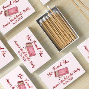May include: White matchboxes with pink and red accents. The boxes have the text "She Found Her" and "Perfect Match" with a graphic of a matchstick. The text "Anna's Bachelorette Party" and the date "11.28.2025" are also on the boxes.