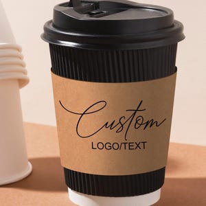 Könnte beinhalten: Ein schwarzer Einweg-Kaffeebecher mit einer braunen Papierhülle. Auf der Hülle steht das Wort "Custom" in Schreibschrift, darunter "LOGO/TEXT". Links ist ein Stapel weißer Becher zu sehen.