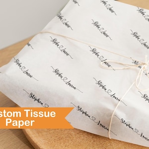 Può includere: Carta velina bianca personalizzata con un motivo a script nero ripetuto con i nomi "Stephen Lauren" e un simbolo a cuore. La carta è avvolta attorno a un oggetto e legata con spago. Le parole "Custom Tissue Paper" sono su uno striscione arancione.