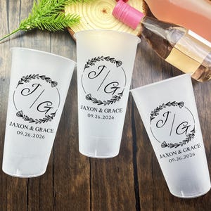 May include: Three frosted plastic cups with a floral wreath design and the initials "J/G" inside a circle. The text "JAXON & GRACE 09.26.2026" is printed below the initials. The cups are on a wooden surface.