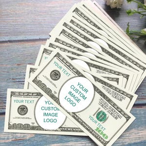 Puede incluir: Un abanico de billetes de 100 dólares estadounidenses con áreas para texto y logotipos personalizados. Los billetes son verdes y negros, con el texto "YOUR TEXT" y "YOUR CUSTOM IMAGE LOGO". Están dispuestos sobre una superficie de madera azul claro.