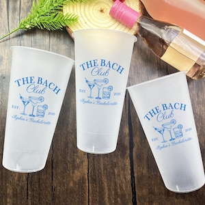 Puede incluir: Tres vasos de plástico esmerilado con el texto "THE BACH Club" en azul, junto con ilustraciones de cócteles y el texto "Sophia's Bachelorette" y el año 2026. Los vasos están sobre una superficie de madera, con una botella de vino rosado al fondo.