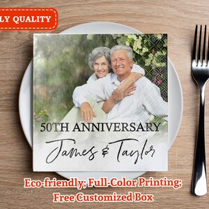 Può includere: Tovagliolo di carta quadrato bianco con un'immagine stampata di una coppia di anziani che si abbracciano, con il testo "50th Anniversary" e i nomi "James & Taylor". Una forchetta nera è su una superficie di legno.