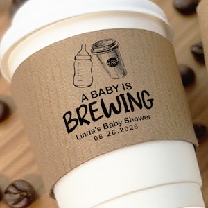 Puede incluir: Una taza de café de papel blanca con una funda marrón. La funda tiene una ilustración de biberón y taza de café, con el texto "A BABY IS BREWING" y "Linda's Baby Shower 08.26.2026". Granos de café esparcidos alrededor de la taza.