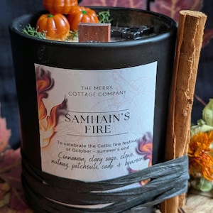 Puede incluir: Una vela negra en un frasco de vidrio con una etiqueta que dice THE MERRY COTTAGE COMPANY SAMHAIN'S FIRE. La parte superior presenta pequeños adornos de cera en forma de calabaza naranja. Una rama de canela está atada al lado con rafia negra.