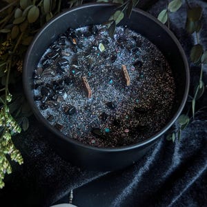 May include: A round black candle container filled with dark wax, topped with scattered black polished stones, fine silver glitter, and small colorful flecks. Two rectangular wooden wicks are positioned in the center of the wax surface.
