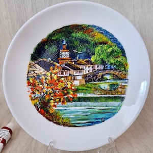 Könnte beinhalten: Handbemalte Keramikplatte mit einer malerischen Szene einer kleinen Stadt mit einem Glockenturm, einer Brücke und einem Fluss. Die Szene ist in leuchtenden Farben gemalt, darunter Grün, Blau, Braun und Rot.