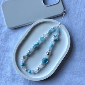 Puede incluir: Un charm de teléfono con cuentas con una variedad de cuentas azules, blancas y plateadas, que incluyen una concha y una cuenta en forma de pez. El charm se muestra en una bandeja ovalada blanca junto a una funda de teléfono gris claro.