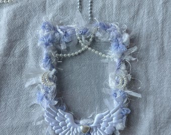 May include: A white and light blue angel wing wreath with a pearl and silver crescent moon embellishment. The wreath is decorated with white and light blue fluffy fabric and has a small white heart charm in the center.