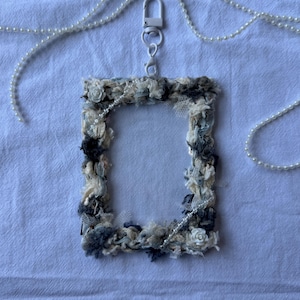 Puede incluir: Un portatarjetas de identificación rectangular con un marco decorativo hecho de tela texturizada en tonos crema, gris y azul. El marco está adornado con pequeñas rosas de tela blanca y una cadena de perlas. Se adjunta un cierre de metal blanco y un collar de perlas.