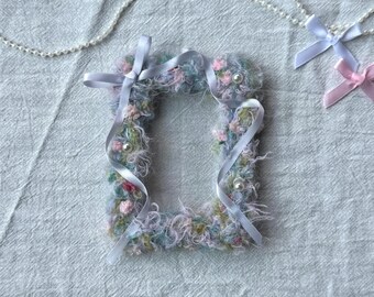 May include: A fluffy, square-shaped decorative item with a central opening, adorned with pastel-colored yarn, small flowers, and white ribbons. Small pearls are interspersed throughout the design. A pearl necklace and two small ribbon bows are also present.