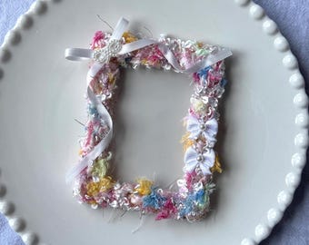 May include: A small, rectangular picture frame with a colorful, textured fabric border. The frame features pink, yellow, and blue threads, accented with white ribbons and small bows. The frame sits on a white, scalloped-edge plate.