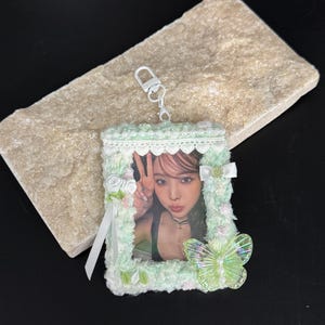 Puede incluir: Un llavero fotográfico verde menta con la foto de una persona. El llavero está decorado con encaje, cintas y una mariposa translúcida. El llavero tiene un cierre plateado.