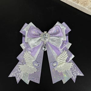 Può includere: Un fiocco decorativo nei toni del lavanda, bianco e argento. Il fiocco presenta un gioiello centrale e un cristallo pendente. Le code del fiocco sono decorate con ali bianche e forme di cuore.