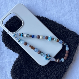 Puede incluir: Un charm de teléfono con cuentas con una variedad de cuentas coloridas, incluyendo azules, marrones y blancas, adjunto a una funda de teléfono blanca. El charm presenta una cuenta en forma de corazón azul. La funda del teléfono descansa sobre una superficie negra texturizada.